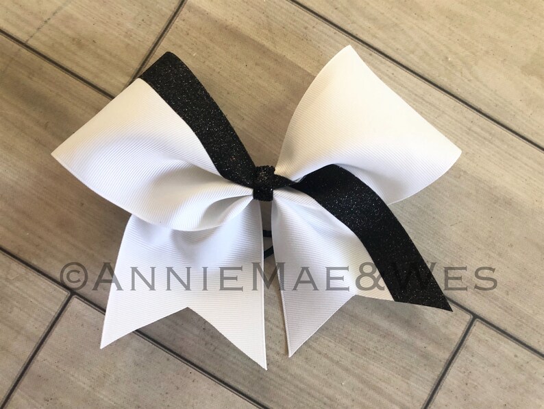 Op de afbeelding: Een witte cheer bow met een zwarte glitterstreep. De strik is vastgebonden met een zwart lint.