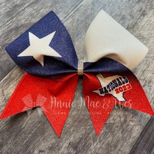 May include: A glittery hair bow featuring the Texas state flag design with a white star on a navy blue background. The bow has red and white accents and the text "Nationals 2023".
