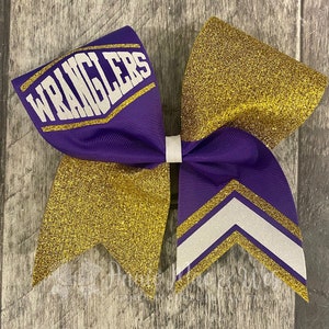 May include: A large purple and gold glitter cheerleading bow. The bow features the word "WRANGLERS" in white block letters on a purple ribbon. Gold glitter accents the bow's design, with white and gold chevron stripes.