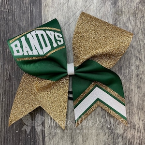 May include: A green and gold cheerleading bow with the word "BANDYS" in white. The bow features gold glitter accents and a white chevron design. The bow is tied with a white ribbon.