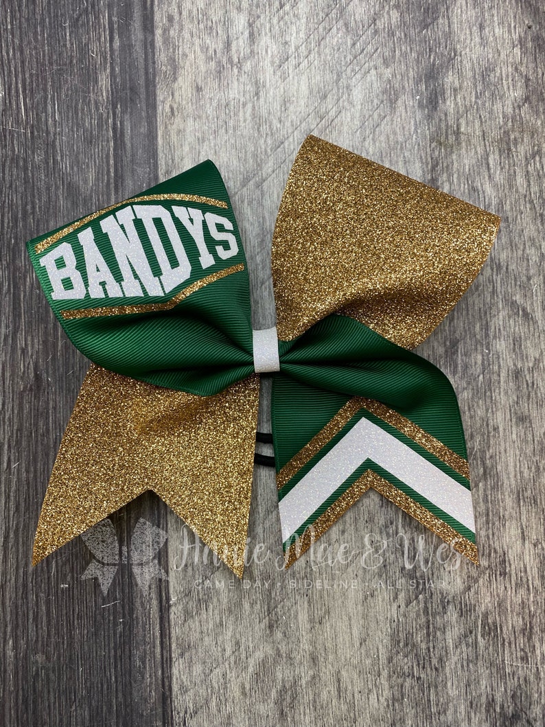 May include: A green and gold cheerleading bow with the word "BANDYS" in white. The bow features gold glitter accents and a white chevron design. The bow is tied with a white ribbon.