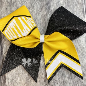 May include: A large cheerleading bow with a yellow and black color scheme. The bow features the word "OUTLAWS" in white block letters on a yellow ribbon. The bow has a glittery black section and a white and black chevron design.
