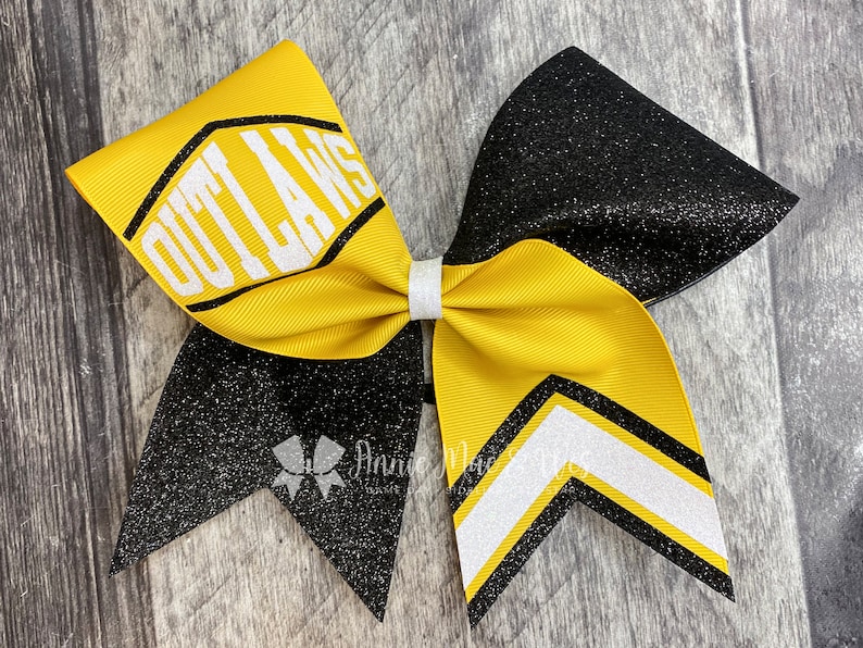 May include: A large cheerleading bow with a yellow and black color scheme. The bow features the word "OUTLAWS" in white block letters on a yellow ribbon. The bow has a glittery black section and a white and black chevron design.