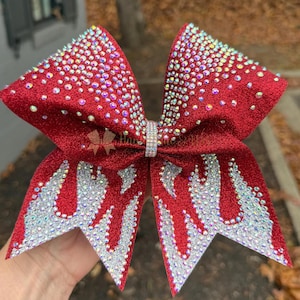 May include: A red glitter cheerleading bow with silver glitter accents and sparkling rhinestones. The bow features a flame-like design on the tails and is embellished with iridescent rhinestones. The centre of the bow is adorned with a rhinestone band.