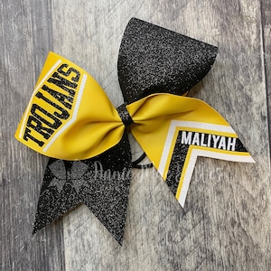 May include: A yellow and black cheer bow with the words "Trojans" and "Maliyah" in white lettering.