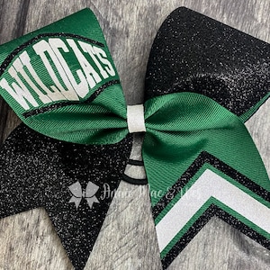 May include: A green and black cheerleading bow with the word "WILDCATS" in white. The bow features a white center knot and a black glitter accent. The bow is designed with a chevron pattern and a black elastic band.