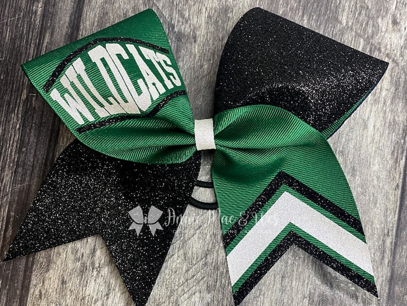 May include: A green and black cheerleading bow with the word "WILDCATS" in white. The bow features a white center knot and a black glitter accent. The bow is designed with a chevron pattern and a black elastic band.
