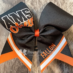 May include: Black and orange cheer bow with white accents. The bow has the text "AMS Cheer" on one side and "FALLON" on the other side.