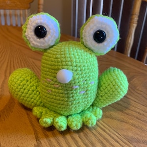 May include: A green crocheted frog with white eyes and black pupils. The frog has a white nose and pink cheeks.
