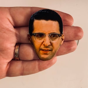 May include: A close-up of a sticker featuring a man with dark hair and glasses. He is wearing a white shirt and has a serious expression on his face.