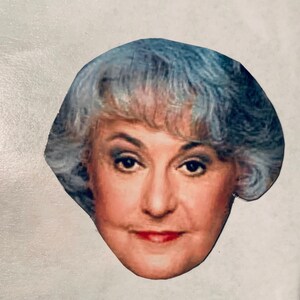 May include: A close-up of a woman's face with short, silver-gray hair. She is looking directly at the camera with a neutral expression.