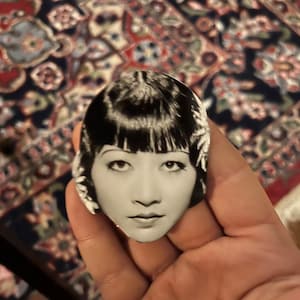 May include: Black and white photo of a woman with short, straight black hair and a serious expression. She is wearing a white dress with a floral pattern.