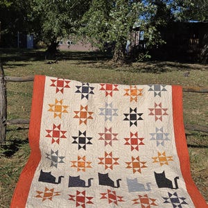 May include: A handmade quilt with a cream-colored background, featuring star patterns in shades of red, orange, black, and gray. The quilt has a rust-colored border and includes cat silhouettes. It is draped over a wooden fence outdoors.