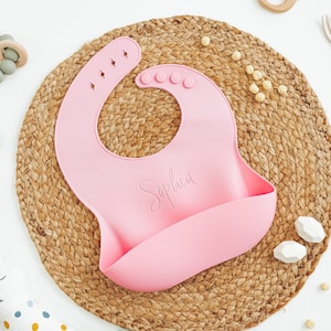 Personalized pink silicone baby bib on woven charger with baby items surrounding by Legacy and Light