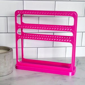 May include: A bright pink earring display stand with multiple rows of small holes for holding earrings. The stand is made of plastic and has a rectangular base. The background is a white tile wall.