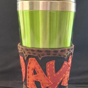 May include: A metallic lime green travel mug with a silver lid, featuring a dark gray felt sleeve. The sleeve is decorated with orange felt letters spelling JAM, attached with visible orange hand-stitched embroidery along the edges.
