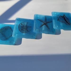 May include: Four blue glass coasters with a white border and black images of a seashell, a starfish, a sand dollar, and a conch shell.