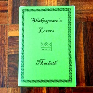 May include: A green paperback book with the titles "Shakespeare's Lovers" and "Macbeth" in a decorative script. The cover has a patterned border and a crown emblem. The book is on a dark wooden surface.