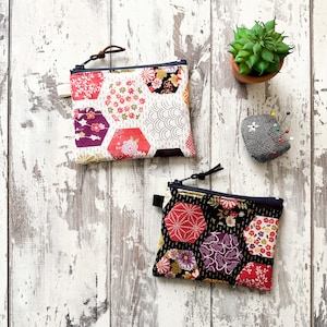 May include: Two fabric zipper pouches with a geometric pattern of hexagons in shades of red, pink, black, and white. The pouches are made of cotton fabric and have a zipper closure.