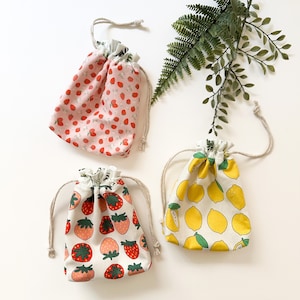 May include: Three drawstring bags with different fruit patterns. One bag has a pink background with white strawberries, another has a white background with red strawberries, and the last bag has a white background with yellow lemons.