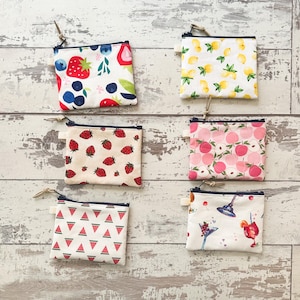 May include: Six small, square zipper pouches with various fruit and cocktail patterns. Designs include strawberries, blueberries, lemons, peaches, watermelon slices, and cocktail glasses. Each pouch has a dark blue zipper and a small leather pull.