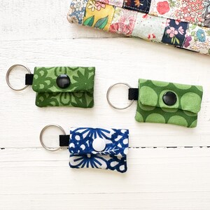 May include: Three keychains with different fabric patterns. Each keychain has a small button and a metal ring. The keychains are green, blue, and white.
