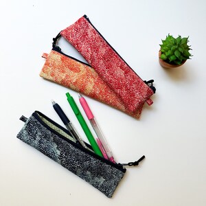 May include: Two fabric pencil cases with zipper closures. The top case is red with white polka dots and the bottom case is blue with white polka dots. The blue case has four pens inside, including a pink, green, and two black pens.