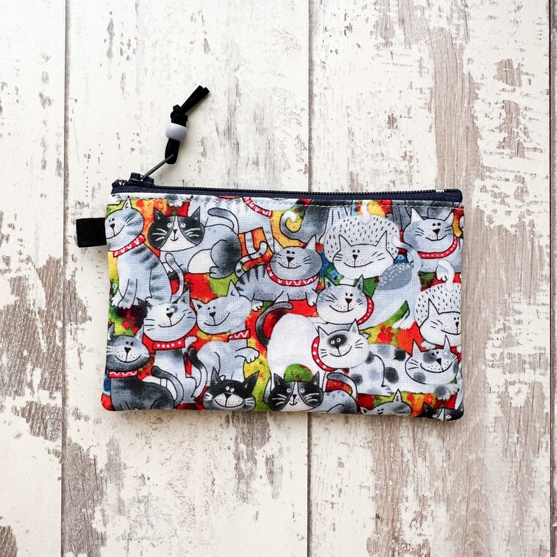 May include: A zippered pouch with a colorful print of cats. The pouch is made of fabric and has a black zipper.