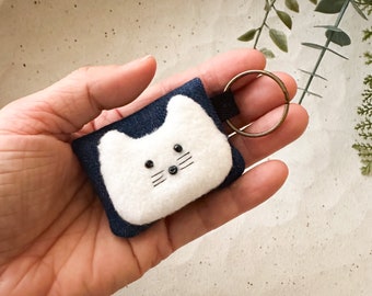 May include: A white felt cat keychain with black eyes and whiskers. The cat is attached to a dark blue felt background and a silver key ring.