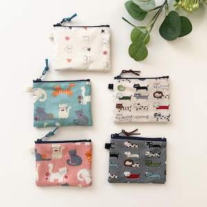 May include: A collection of small, square coin purses with zipper closures. Each purse features a unique cat-themed pattern in various colors, including white, blue, pink, and gray. The designs showcase playful cat illustrations.