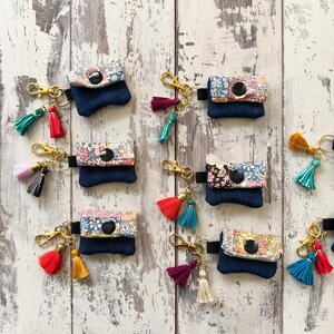May include: A collection of eight small denim pouches with floral fabric accents and a black button. Each pouch is attached to a gold key chain with a tassel. The tassels are in various colors, including red, pink, blue, and yellow.