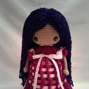 May include: A crocheted doll with long dark blue hair wearing a burgundy dress with pink ribbon and white hearts.