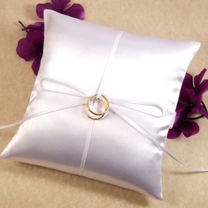 May include: White satin pillow with a white satin ribbon tied in a bow. Two gold wedding bands are placed on top of the bow.