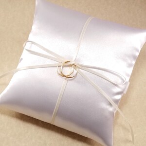 White satin ring pillow on a beige background. It has a simple bow with imitation gold rings tied at the center.