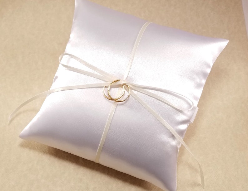 White satin ring pillow on a beige background. It has a simple bow with imitation gold rings tied at the center.