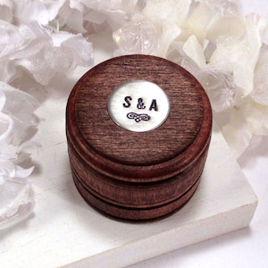 May include: A dark brown wooden ring box with a silver metal lid. The lid has a circular engraving that reads "S&A" with a decorative design.