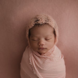 May include: A newborn baby is swaddled in a pink fabric and wearing a pink crocheted hat. The baby is sleeping peacefully with their eyes closed.