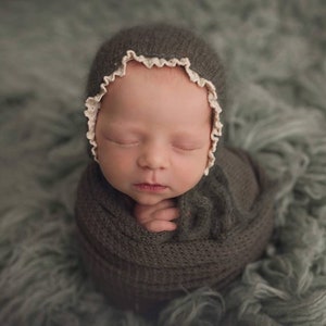 May include: A newborn baby is swaddled in a dark gray knitted wrap and wearing a matching knitted hat with a white lace trim. The baby is sleeping peacefully with their eyes closed.
