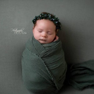 May include: A newborn baby girl is sleeping peacefully, wrapped in a soft green swaddle. She is wearing a green and white floral crown.