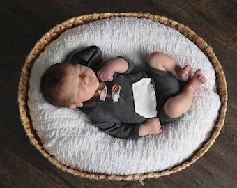 Könnte beinhalten: Ein neugeborenes Baby schläft in einem geflochtenen Korb, trägt einen grauen Kapuzen-Strampler mit Holzknöpfen. Das Baby liegt auf einer weißen, flauschigen Decke.