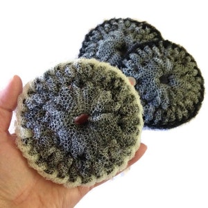 May include: Three black and white crocheted scrubbers, two are smaller and one is larger. The scrubbers are made of a soft, textured material and have a hole in the center.