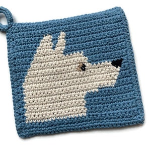 May include: Crocheted blue pot holder with a white dog design. The dog's face is stitched in white yarn with black eyes and nose.