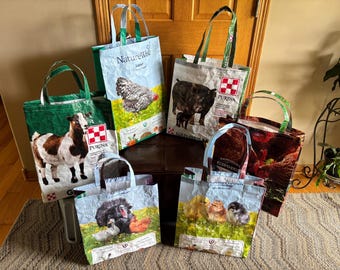Puede incluir: Una colección de seis bolsas de tela reutilizables hechas de sacos de pienso reciclados. Cada bolsa presenta impresiones fotográficas coloridas de animales de granja, incluyendo una cabra, un pavo, pollitos y un cerdo, con marcas Purina y NatureWise.