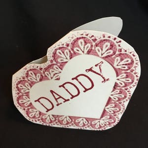 May include: A white heart-shaped card with a pink lace border. The word "DADDY" is printed in red on the card.