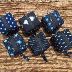 May include: Six small, square fabric pouches with zippers, each featuring distinct patterns including blue and white geometric shapes, black and white abstract designs, and dark blue with white polka dots, arranged in a cluster.