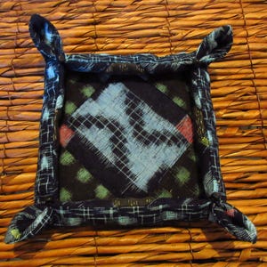 May include: Square fabric tray with a dark blue and white patterned exterior and a light blue and black patterned interior. The tray has four corner accents. The tray is on a brown woven surface.