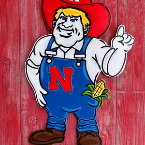 May include: A wooden cutout of a cartoon farmer wearing a red hat with a white "N" on it, blue overalls, and a white shirt. The farmer is holding a corn cob in his right hand and pointing with his left hand. The cutout is painted in bright colors and has a sign at the bottom that reads "Knock Knocks Door Hangs and More".