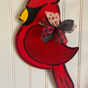 May include: A red cardinal wooden door hanger with a black outline. The cardinal has a yellow beak and a black eye. The cardinal is decorated with a bow made of red and black plaid fabric, brown burlap fabric with white polka dots, and black fabric with white polka dots.