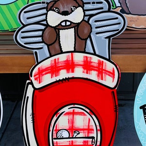 May include: A red and white plaid golf bag with a brown and white beaver holding golf clubs. The bag has a golf ball and a tee inside.