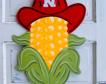 May include: A red and white corn on the cob door hanger with a red cowboy hat with a white "N" on it. The door hanger is decorated with a red and white gingham bow. The sign at the bottom reads "Knock Knocks Door Hangs and More".